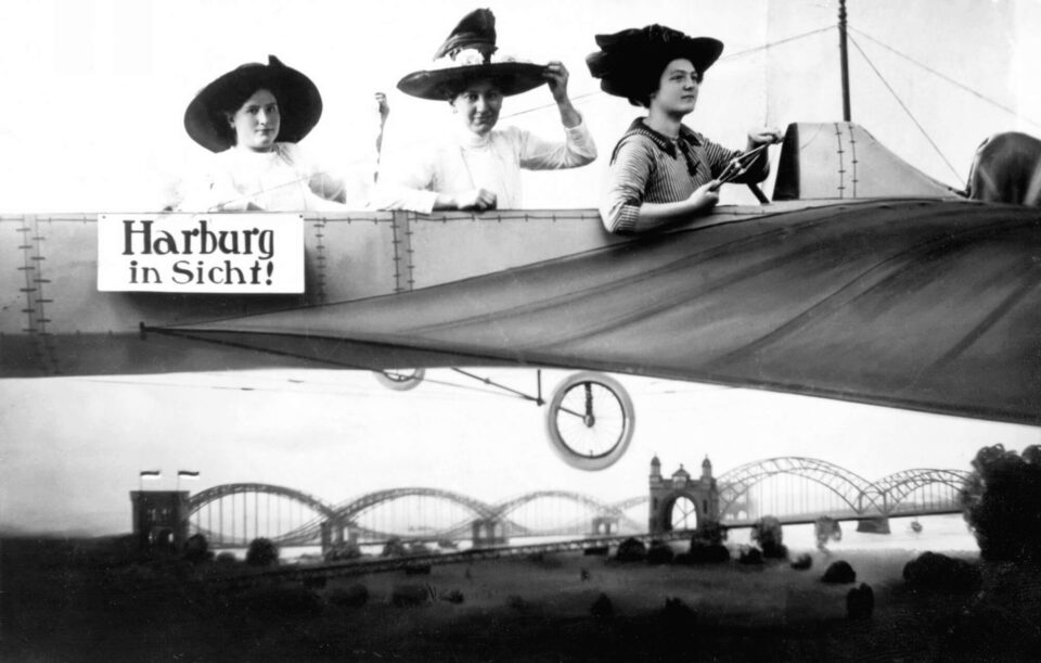 Drei Frauen posieren in einer Flugzeug-Attrappe, das beschriftet ist mit "Harburg in Sicht!". Im Hintergrund sind die Brücken über die Süderelbe abgebildet.