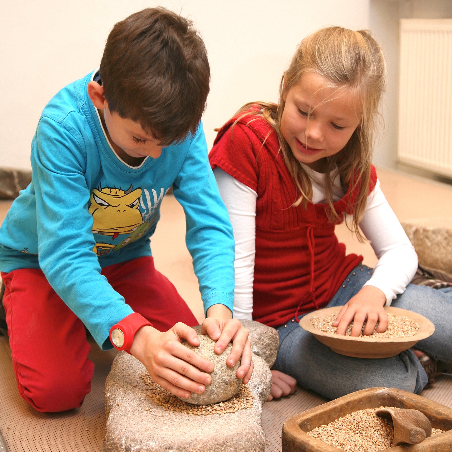 Zwei Kinder mahlen auf steinernen Mahlsteinen Korn zu Mehl.