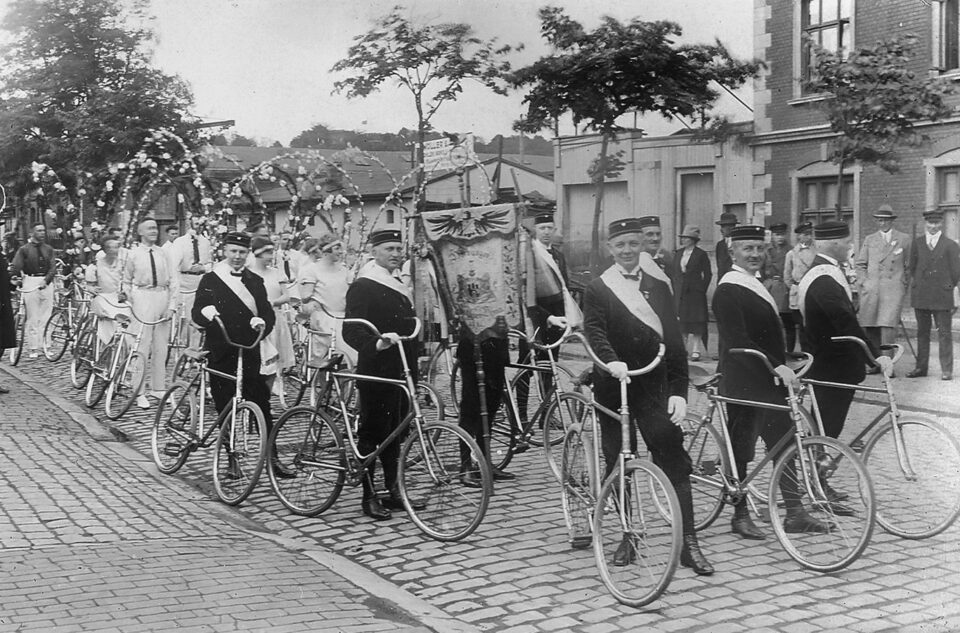 Ein Festumzug. Eine festlich gekleidete und geschmückte Gruppe schiebt Fahrräder und trägt eine Vereinfahne.
