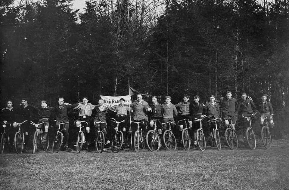 Vor einem Waldrand aufgereiht: eine Mannschaft auf ihren Fahrrädern sitzen, einander die Arme auf die Schultern gelegt zum Mannschaftsfoto